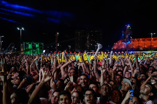 Plateia durante o Rock in Rio de 2015, com braços levantados e luzes azuis verdes e amarelas ao fundo.